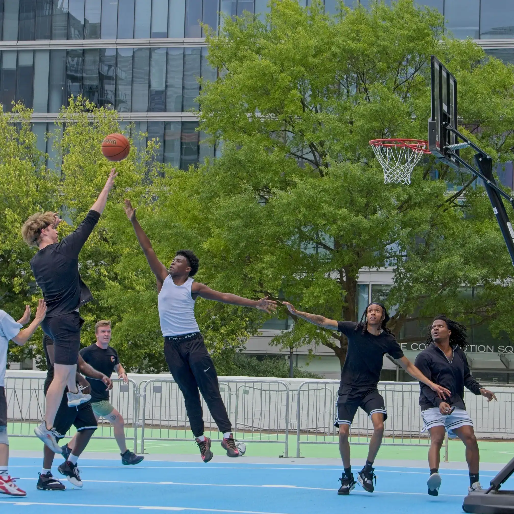 A player shoots over a leaping defender on a bright blue pop-up basketball court ringed by office buildings