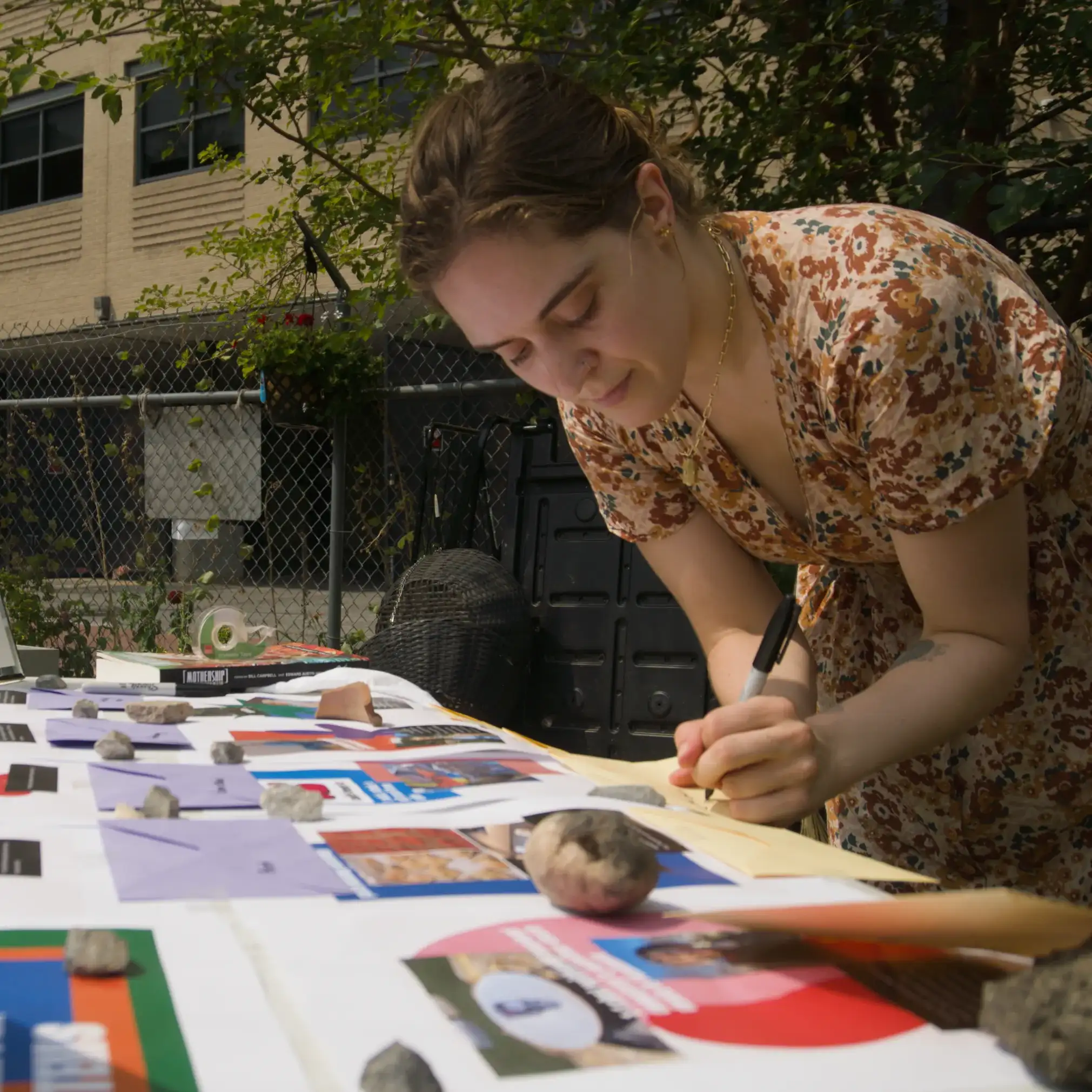 A woman in a floral dress writing a note at a table spread with posters, envelopes and stones