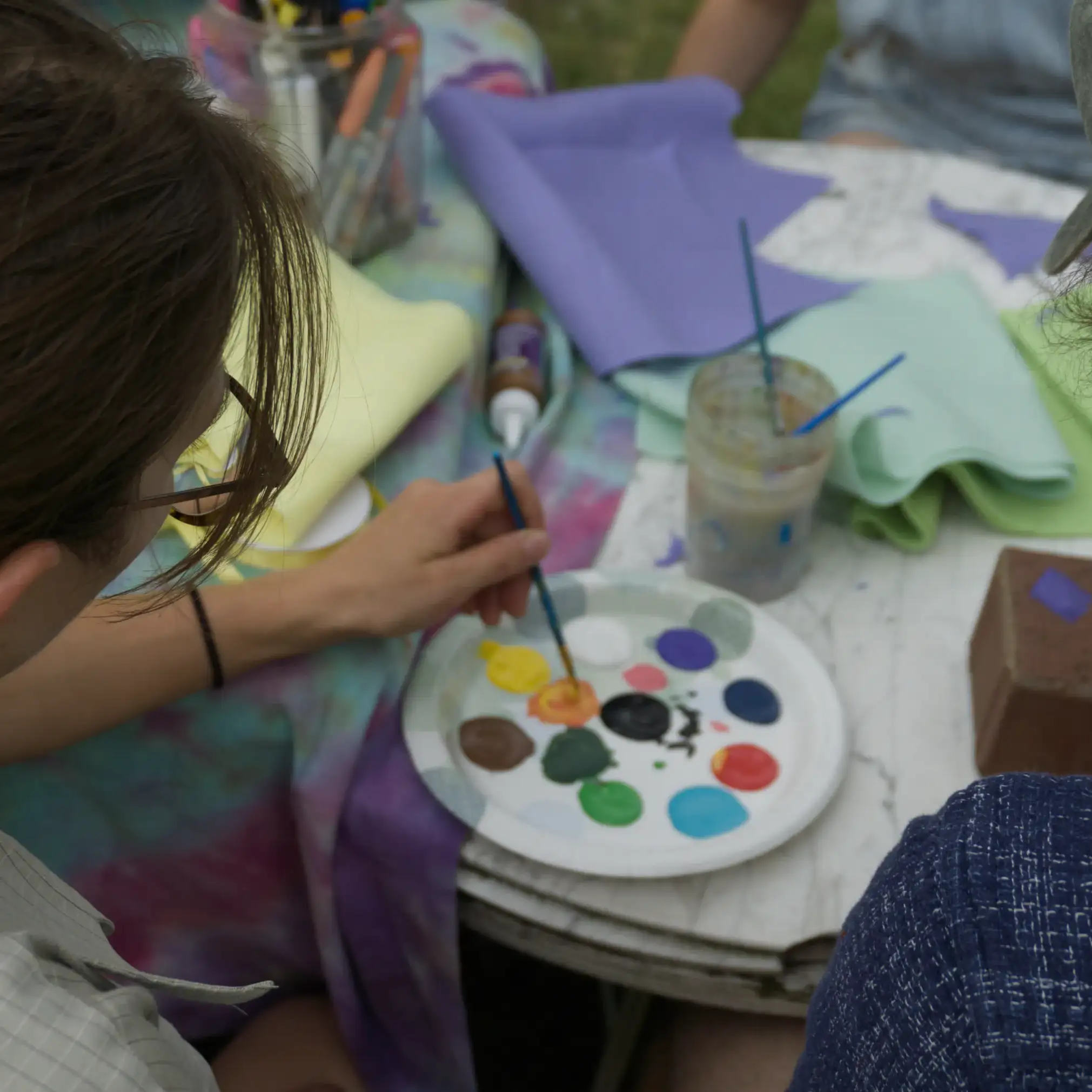 Two people painting at a garden table, one dipping a brush into a paper-plate palette